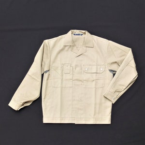 Puede incluir: Una camisa de trabajo beige de manga larga, dos bolsillos en el pecho y cierre de botones en la parte delantera. La camisa tiene cuello y una etiqueta en el interior del cuello que dice "M.T.M."