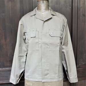 Puede incluir: Una chaqueta de trabajo beige de manga larga, con cierre de botones y dos bolsillos grandes en el pecho.