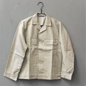 Puede incluir: Una chaqueta de trabajo beige con cierre de botones, dos bolsillos en el pecho y mangas largas. La chaqueta tiene una etiqueta que dice "M.M."