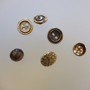 Op de afbeelding: Assortiment van zes goudkleurige knopen met verschillende ontwerpen, waaronder een swirl, een ontwerp met twee gaten, een ontwerp met vier gaten en een textuurontwerp.