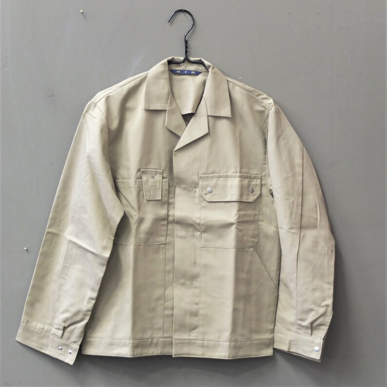 Puede incluir: Una chaqueta de trabajo beige con cierre de botones, dos bolsillos en el pecho y mangas largas. La chaqueta tiene una etiqueta que dice "M.M."