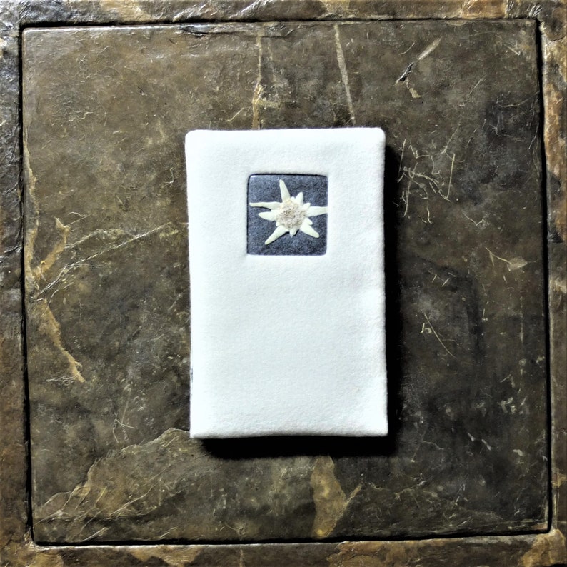 Puede incluir: Un cuaderno de fieltro blanco con un peque&ntilde;o parche cuadrado azul que presenta una flor de edelweiss blanca.