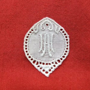 May include: White embroidered lace applique with a scalloped edge and a letter "M" in the centre, on a red fabric background.