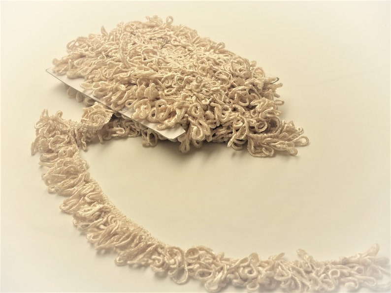 May include: A close-up of a beige, curly, decorative trim. The trim is made of a soft, woven fabric and has a delicate, fringed texture.