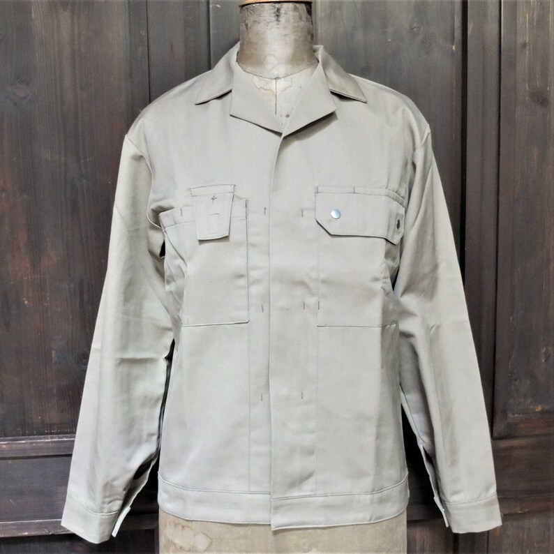 Puede incluir: Una chaqueta de trabajo beige de manga larga, con cierre de botones y dos bolsillos grandes en el pecho.