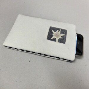Puede incluir: Funda de tel&eacute;fono de fieltro blanco con ribete a cuadros blanco y negro. Un peque&ntilde;o cuadrado gris con una flor de edelweiss blanca est&aacute; centrado en la funda.
