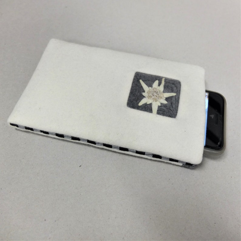 Puede incluir: Funda de tel&eacute;fono de fieltro blanco con ribete a cuadros blanco y negro. Un peque&ntilde;o cuadrado gris con una flor de edelweiss blanca est&aacute; centrado en la funda.
