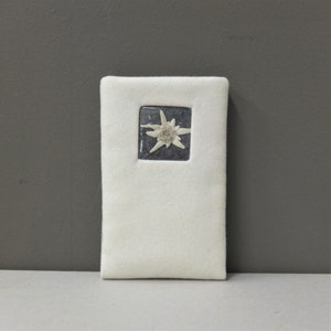 Puede incluir: Un marcap&aacute;ginas de fieltro blanco con una imagen enmarcada de una flor de edelweiss blanca sobre un fondo gris oscuro.