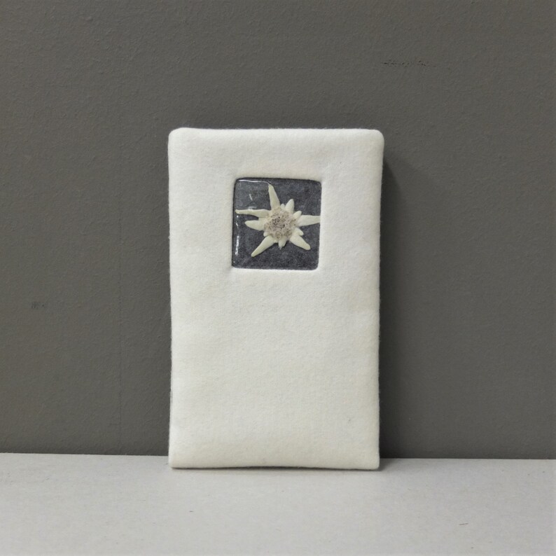Puede incluir: Un marcap&aacute;ginas de fieltro blanco con una imagen enmarcada de una flor de edelweiss blanca sobre un fondo gris oscuro.