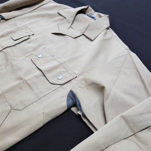 Puede incluir: Una camisa de trabajo de color beige con cuello abotonado y un panel de malla en el codo. La camisa tiene dos bolsillos grandes en la parte delantera.