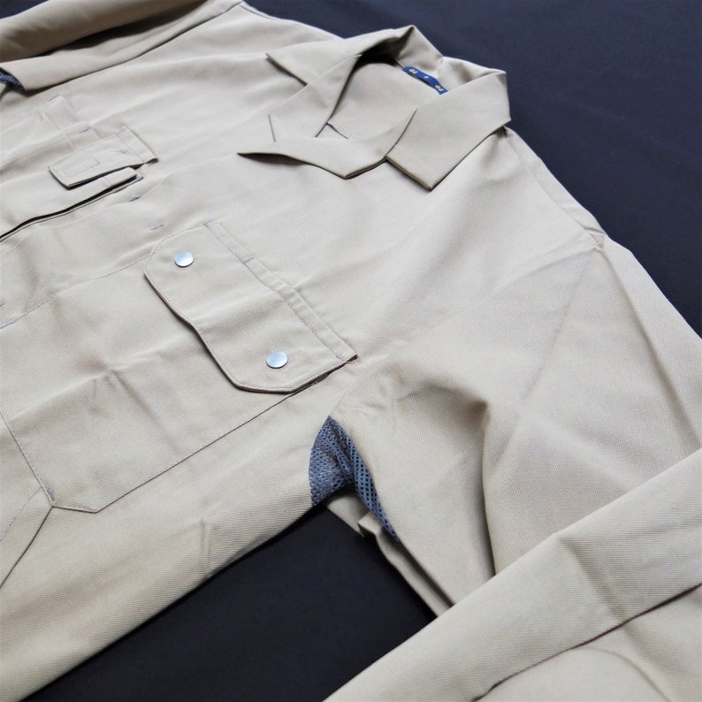 Puede incluir: Una camisa de trabajo de color beige con cuello abotonado y un panel de malla en el codo. La camisa tiene dos bolsillos grandes en la parte delantera.