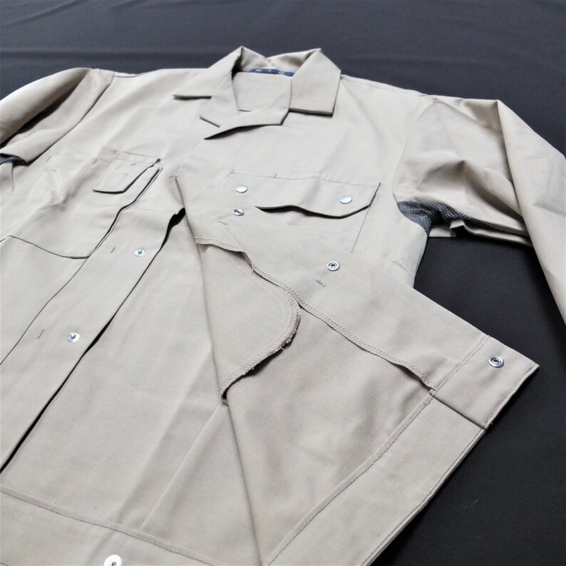 Puede incluir: Una camisa de trabajo beige con botones y un solo bolsillo en el pecho. La camisa est&aacute; hecha de una tela duradera y tiene un corte holgado.
