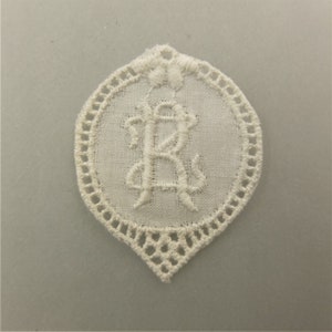 Könnte beinhalten: Weiße Spitzentischdecke mit einem zarten gestickten Buchstaben "R" in der Mitte.