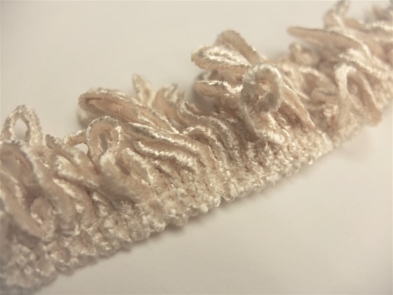 May include: Close-up of a cream-colored fabric trim with a ruffled texture. The trim is made of soft, delicate fibers and has a slightly uneven surface.