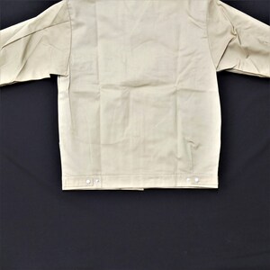 Puede incluir: Una chaqueta de trabajo beige con cierre de botones y dos bolsillos. La chaqueta tiene un dise&ntilde;o sencillo y est&aacute; hecha de un tejido resistente.