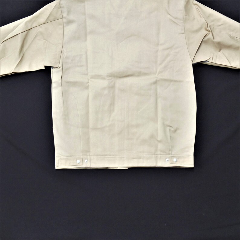 Puede incluir: Una chaqueta de trabajo beige con cierre de botones y dos bolsillos. La chaqueta tiene un dise&ntilde;o sencillo y est&aacute; hecha de un tejido resistente.