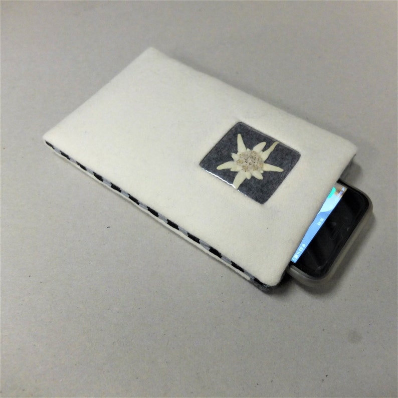 Puede incluir: Funda para tel&eacute;fono de fieltro blanco con ribete a cuadros blanco y negro. Un cuadrado de pl&aacute;stico transparente con una flor de edelweiss blanca est&aacute; centrado en la funda. Un tel&eacute;fono inteligente negro est&aacute; parcialmente insertado en la funda.
