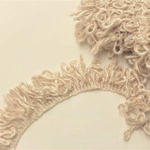 May include: Cream-colored fabric trim with a fringe design. The trim is made of soft, looped fabric and has a delicate, feminine look.