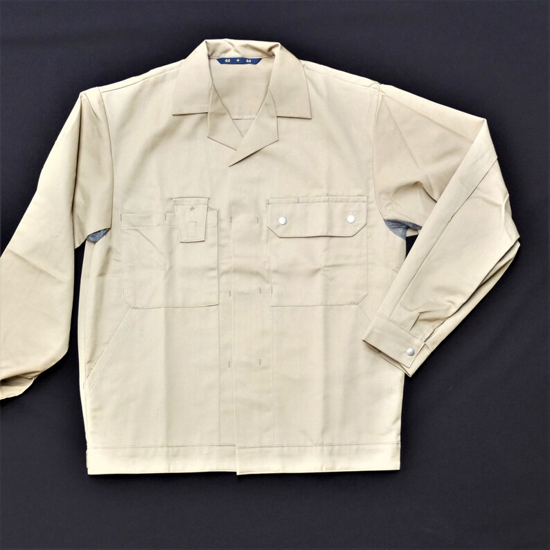 Puede incluir: Una camisa de trabajo beige de manga larga, cuello abotonado y dos bolsillos en el pecho. La camisa tiene una etiqueta en la parte delantera que dice "M-L".