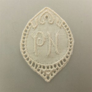 Könnte beinhalten: Weißer Baumwollstoff mit zarter Spitzenborte und den gestickten Buchstaben "PN" in der Mitte.
