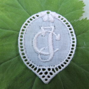 May include: White lace oval with a white embroidered letter "J" in the centre. The lace has a scalloped edge.