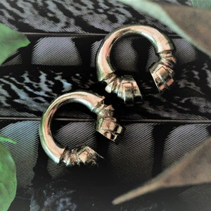 May include: Two gold-colored metal hoop earrings with a textured, geometric design. The earrings are on a black and white patterned surface.