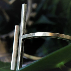 May include: A silver ring with a simple, geometric design. The ring is made of two thin, rectangular bars that intersect at a right angle. The bars are smooth and polished.