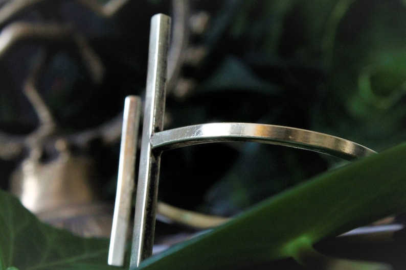 May include: A silver ring with a simple, geometric design. The ring is made of two thin, rectangular bars that intersect at a right angle. The bars are smooth and polished.