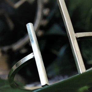 May include: A silver metal bangle bracelet with a simple geometric design. The bracelet is made of two straight bars that intersect at right angles.