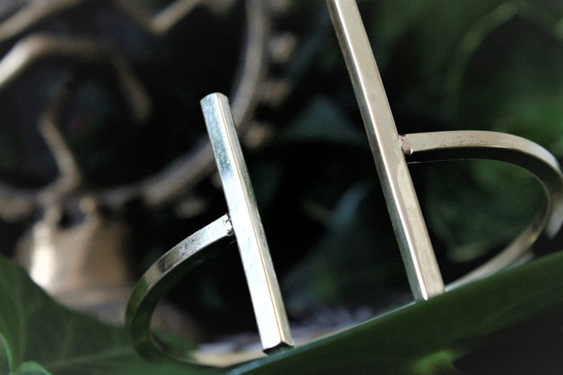 May include: A silver metal bangle bracelet with a simple geometric design. The bracelet is made of two straight bars that intersect at right angles.