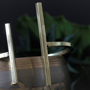 May include: A gold metal bangle bracelet with two vertical bars. The bracelet is open and has a simple design.