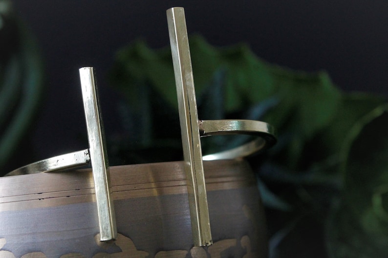 May include: A gold metal bangle bracelet with two vertical bars. The bracelet is open and has a simple design.
