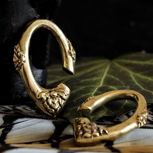 Può includere: Due orecchini a cerchio in metallo color oro con intricati disegni floreali. Gli orecchini sono su uno sfondo di piume e una foglia verde.