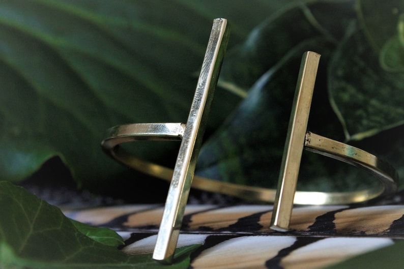 May include: A gold-toned metal ring with a thick, straight bar design. The ring is open and adjustable.