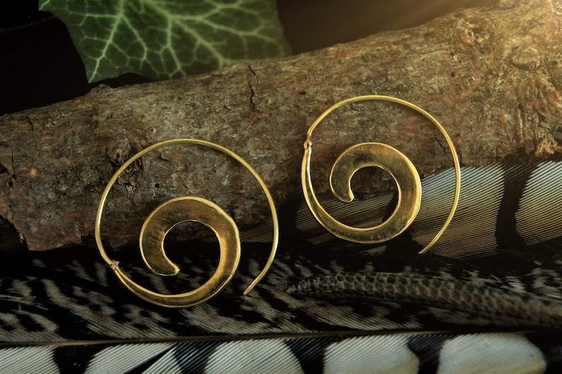 May include: A pair of gold-toned hoop earrings with a spiral design. The earrings are on a wooden surface with a feather background.