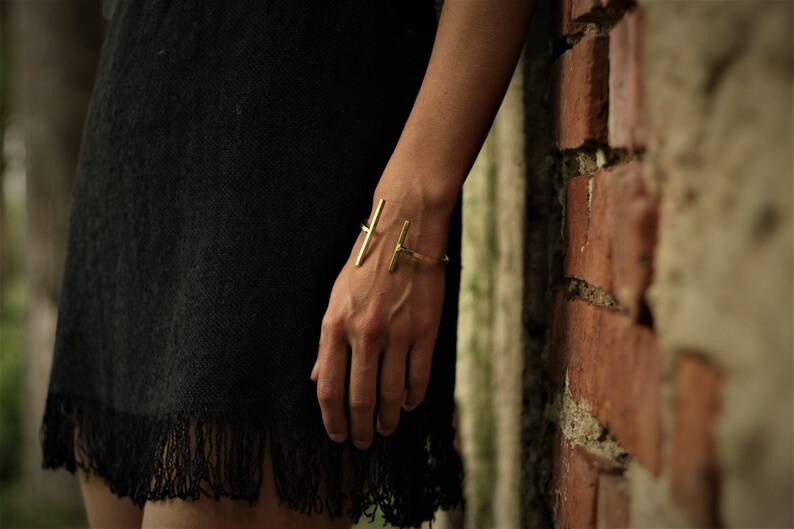 May include: A gold metal bracelet with a simple, minimalist design. The bracelet is made of two thin, straight bars that intersect at a right angle.