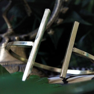 May include: A pair of gold-toned metal bracelets with a simple, minimalist design. Each bracelet features a straight, flat bar that intersects with a circular band.
