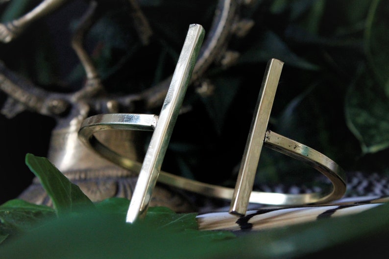 May include: A pair of gold-toned metal bracelets with a simple, minimalist design. Each bracelet features a straight, flat bar that intersects with a circular band.