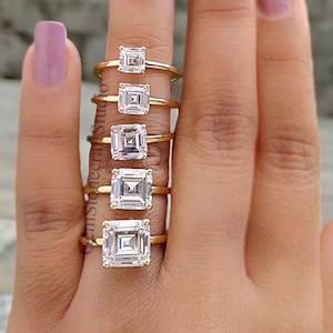 May include: A set of seven gold rings with square-shaped clear gemstones of varying sizes. The rings are stacked on a finger.