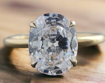 May include: A gold engagement ring with a large, oval-cut diamond. The diamond is clear and multifaceted, set with four prongs. The ring band is a simple, polished gold. The ring is displayed on a wooden surface.