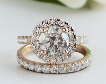 May include: A rose gold engagement ring set with a large, round, clear center stone surrounded by smaller stones. The band of the engagement ring and the accompanying wedding band are also adorned with small, clear stones. The rings are set against a soft, white background.