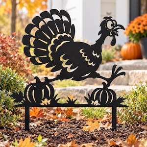 Puede incluir: Un adorno de jardín de metal negro con la silueta de un pavo de cola desplegada y ojos muy abiertos, situado sobre dos calabazas y hojas de otoño, sostenido por dos estacas verticales para insertar en el suelo.