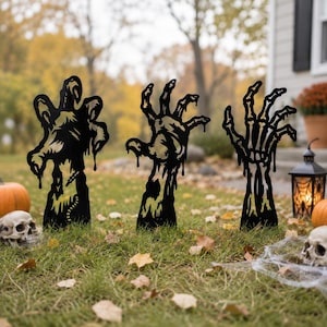 Peut inclure: Trois silhouettes en métal noir représentant des mains qui sortent du sol. Les motifs incluent une main texturée avec des détails dégoulinants, une main en forme de griffe aux doigts pointus et une main squelettique avec des os apparents.