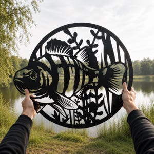 May include: A circular black metal wall art piece featuring a detailed cutout silhouette of a striped perch fish swimming among aquatic plants. The design is enclosed within a solid circular frame, showcasing intricate lines and negative space.