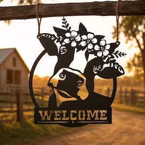 May include: A black metal wall hanging sign featuring a cutout silhouette of a cow wearing a floral crown. The circular design includes the word WELCOME in capital letters at the bottom and is suspended by two pieces of brown twine.