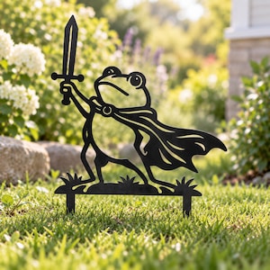 May include: A black metal garden stake featuring a silhouette of a frog wearing a cape and holding a sword aloft. The frog stands on a small base with grass-like details, designed to be inserted into the ground as a decorative outdoor accent.