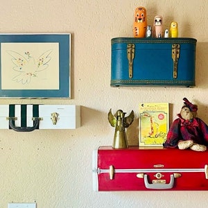May include: A white and black striped suitcase with gold hardware sits on a wall shelf. Above it is a framed drawing of a white dove with colorful flowers. On the shelf below, a red suitcase with gold hardware is decorated with a teddy bear, a book titled "The Washington Rabbit", and a gold angel figurine.