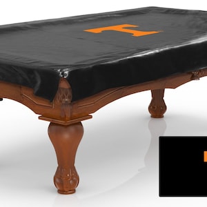 May include: A black pool table cover with an orange "T" logo. The cover is draped over a wooden pool table with ornate legs and decorative pockets. A smaller image of the logo is in the bottom right corner.