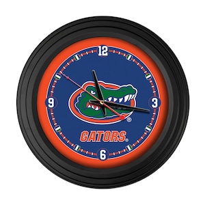 May include: Round black wall clock with a blue face featuring the University of Florida Gators logo. The clock has a black bezel, orange inner ring, and white numerals at the 3, 6, 9, and 12 o'clock positions. The clock hands are black.