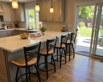 May include: A modern kitchen with a light-colored countertop island and black bar stools with wooden seats. The kitchen features light-colored cabinets, pendant lights, and a view of a backyard through a sliding glass door.